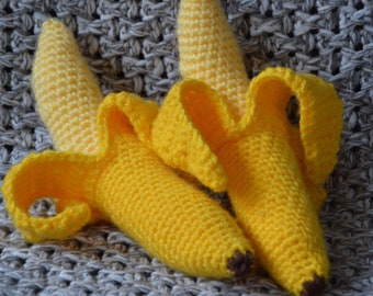 May include: Two crocheted banana toys, one with the peel partially open, are lying on a grey knitted surface.