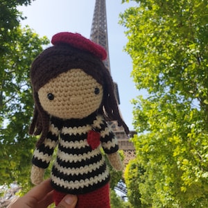 May include: A crocheted doll wearing a red beret and a black and white striped shirt. The doll is holding a red heart and is standing in front of the Eiffel Tower.