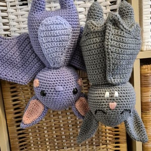 May include: Two crocheted stuffed bats, one purple and one gray, hanging from a wicker basket. The purple bat has pink ears and black eyes. The gray bat has white eyes and a pink nose.