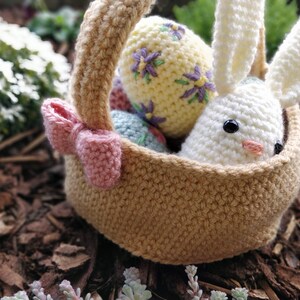 May include: A crocheted bunny rabbit sits in a brown crocheted basket with a pink bow. The basket is filled with colorful crocheted Easter eggs.