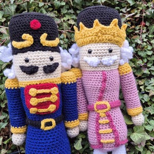 May include: Two crocheted nutcracker dolls, one in blue and gold and the other in pink and gold. The dolls are standing in front of a green leafy background.