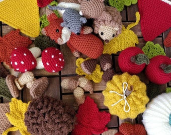 May include: A collection of crocheted woodland animal and nature-themed ornaments, including a fox, a squirrel, a hedgehog, mushrooms, acorns, and leaves. The ornaments are made in various shades of brown, red, yellow, and green.