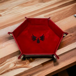 Könnte beinhalten: Rotes Filz-Würfeltablett mit einem schwarzen geflügelten Tropfen-Design. Das Tablett ist sechseckig und hat einen schwarzen Lederrahmen.