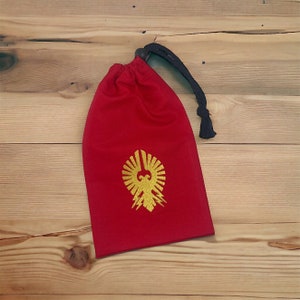 May include: Red drawstring bag with a gold embroidered eagle design. The bag has a black drawstring and a wooden toggle.