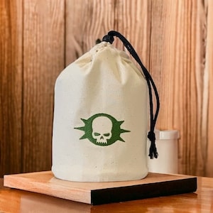 Pode incluir: Uma bolsa de cordão branco com um design de caveira e ossos cruzados verdes. A bolsa está sobre uma superfície de madeira.