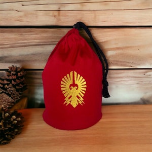 May include: Red drawstring bag with a gold embroidered design of a stylized bird with wings spread. The bag has a black drawstring and tassel.