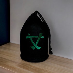 Puede incluir: Una bolsa de cordón negro con un símbolo bordado en verde. El símbolo es una "X" estilizada con dos líneas que se cruzan en el centro.