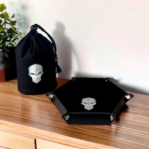 May include: Black dice bag and tray with a skull design. The bag is made of fabric and has a drawstring closure. The tray is made of leather and has a hexagonal shape.