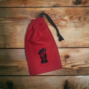 May include: A red drawstring bag with a black embroidered design of a hand holding a bunch of keys. The bag is made of a soft, cotton fabric and has a black drawstring closure.