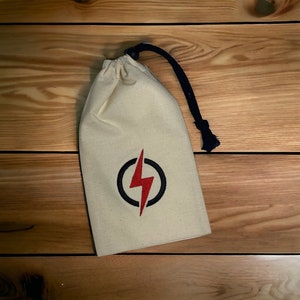 Pode incluir: Uma bolsa de tecido branco com um cordão de ajuste preto e um raio vermelho em um círculo preto na frente.