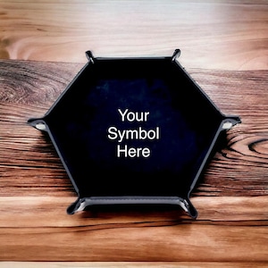 Op de afbeelding: Een zwarte, zeshoekige tray met de tekst "Your Symbol Here" in het wit. De tray heeft een donkerblauwe binnenkant en staat op een houten achtergrond. De tray heeft vier hoekaccenten.