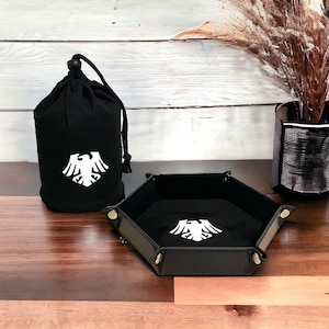 Può includere: Borsa da dado nera e vassoio con un logo di aquila bianca. La borsa è realizzata in tessuto e ha una chiusura con coulisse. Il vassoio è realizzato in pelle e ha una forma esagonale.
