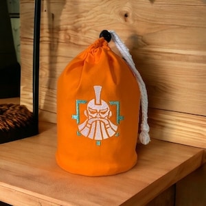 May include: Orange drawstring bag with a white embroidered design of a person with a beard and a crown. The design is framed in a green square.