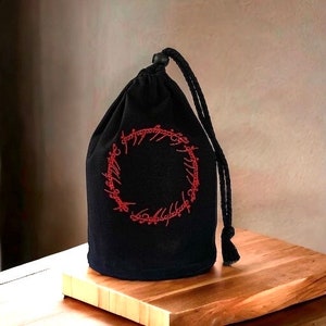 Pode incluir: Saco de cordão preto com um anel e uma inscrição bordados em vermelho. A inscrição diz "Um Anel para governá-los a todos, Um Anel para encontrá-los, Um Anel para trazê-los a todos e nas trevas prendê-los."