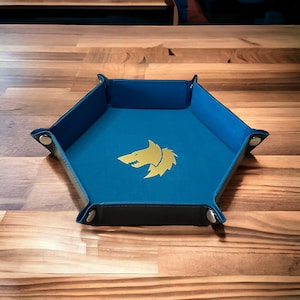 Puede incluir: Una bandeja hexagonal azul para dados con un logotipo de lobo dorado. La bandeja está hecha de cuero y tiene un forro de fieltro.