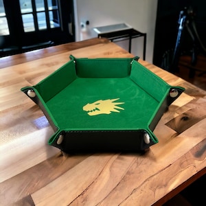 May include: A green felt-lined hexagonal dice tray with a gold dragon silhouette. The tray has a black leather exterior and silver metal accents.