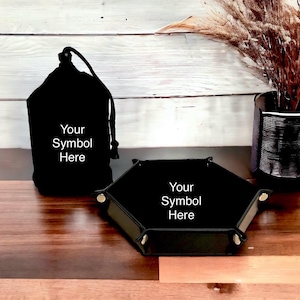 Può includere: Vassoio da gioco a forma esagonale in pelle nera con dettagli dorati. Il vassoio ha la scritta "Your Symbol Here" stampata. È presente anche un sacchetto da gioco in velluto nero con coulisse.