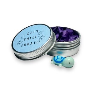May include: A small, silver tin with a blue lid that says "Let's Shell-ebrate!" with fireworks around the text. The tin is open and a small, light blue glass turtle figurine is sitting in front of it.