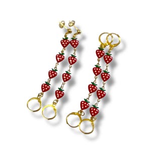 May include: A pair of gold-coloured dangle earrings. Each earring features a chain of red strawberries with white seeds and green tops. The earrings have a hoop closure. The earrings are a fun accessory.
