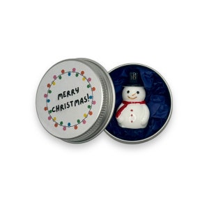 May include: A small, white snowman figurine with a black top hat and red scarf is inside a silver tin with a white label that says "Merry Christmas!" with colourful lights around the text.