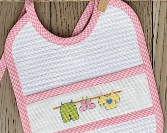 May include: A white bib with a pink gingham trim and a pocket. The pocket has an embroidered design of clothes hanging on a clothesline.