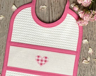 May include: A white baby bib with a pink trim and strap. The bib features a waffle-weave texture and a horizontal pink band with a cross-stitched heart. Dried rose petals are scattered around the bib.
