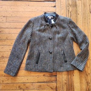 May include: A black and white tweed blazer with a black faux leather collar and trim. The blazer has two pockets with black faux leather trim and two black buttons.