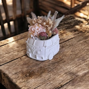 Könnte beinhalten: Ein weißer Keramik-Blumentopf in Form eines Oldtimer-Vans, gefüllt mit getrockneten Blumen in Rosa, Beige und Braun. Der Topf steht auf einer rustikalen Holzoberfläche und erzeugt eine charmante, böhmische Ästhetik. Die Anordnung umfasst Pampasgras und andere strukturierte Blüten.