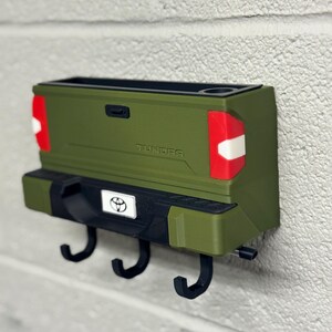 Puede incluir: Un llavero de pared con forma de la parte trasera de una camioneta Toyota Tundra verde. Cuenta con tres ganchos negros, un emblema de Toyota y la palabra "TUNDRA". El dise&ntilde;o incluye luces traseras rojas y blancas y un portavasos.