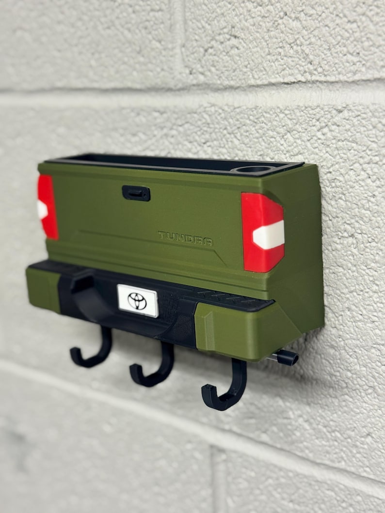 Puede incluir: Un llavero de pared con forma de la parte trasera de una camioneta Toyota Tundra verde. Cuenta con tres ganchos negros, un emblema de Toyota y la palabra "TUNDRA". El dise&ntilde;o incluye luces traseras rojas y blancas y un portavasos.