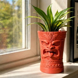May include: A red tiki-style planter pot with a detailed carved face design, holding a green and yellow plant. The planter is cylindrical and sits on a windowsill, with a window and natural light in the background.