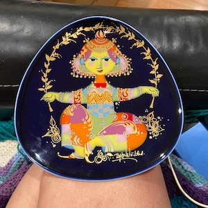 May include: A navy blue, triangular decorative plate with a colourful illustration of a seated figure with outstretched arms. The figure has gold accents and is in orange, purple, and yellow. The plate has a gold border. The plate is approximately 20 cm in diameter.