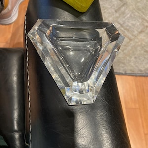 May include: A clear, faceted crystal ashtray in a diamond shape. The ashtray has a triangular bowl and is made of thick, transparent glass. The edges are sharply cut, reflecting light.