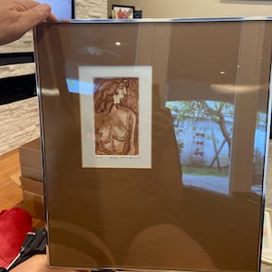 Op de afbeelding: Een ingelijst kunstwerk met een sepia-getinte ets van een naakte figuur. Het kunstwerk wordt getoond in een zilverkleurige lijst, tegen een bruine passe-partout. De afbeelding toont een zijaanzicht van de figuur, met zichtbare details van de torso en het bovenlichaam.