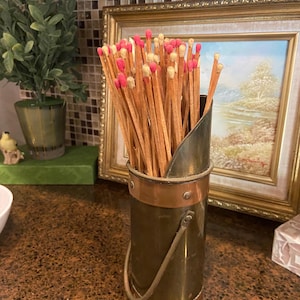 May include: A vintage brass match holder filled with wooden matches. The matches have pink and cream-colored tips. The holder has copper accents and a handle. A framed painting and a small potted plant are in the background.