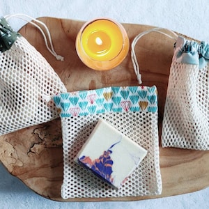 Peut inclure: Plusieurs sacs en filet réutilisables avec des fermetures à cordon et des motifs colorés. Un pain de savon aux motifs colorés est placé dans l'un des sacs. Une bougie allumée est posée à côté sur une surface en bois.
