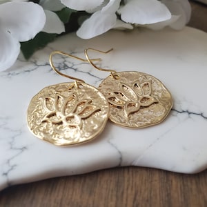 Puede incluir: Pendientes de color dorado con un dise&ntilde;o de flor de loto. Los pendientes son redondos y tienen una superficie texturizada.