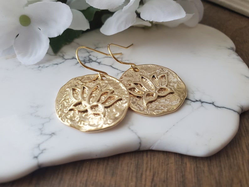 Puede incluir: Pendientes de color dorado con un dise&ntilde;o de flor de loto. Los pendientes son redondos y tienen una superficie texturizada.