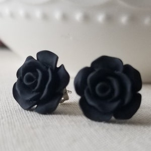 May include: A pair of black rose-shaped stud earrings. The earrings are made of a matte black material and feature detailed petals. The earrings are set against a neutral background, highlighting their dark color and floral design.