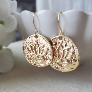 Puede incluir: Pendientes de tono dorado con un dise&ntilde;o redondo texturizado que presenta un motivo de flor de loto en relieve.