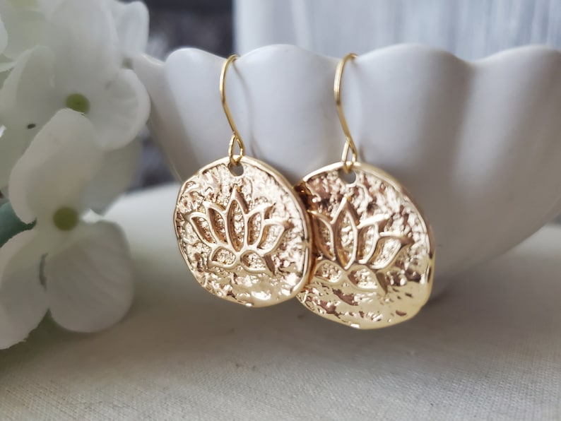 Puede incluir: Pendientes de tono dorado con un dise&ntilde;o redondo texturizado que presenta un motivo de flor de loto en relieve.