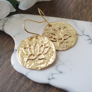 Puede incluir: Pendientes de color dorado con un dise&ntilde;o de flor de loto. Los pendientes son redondos y tienen una textura martillada.