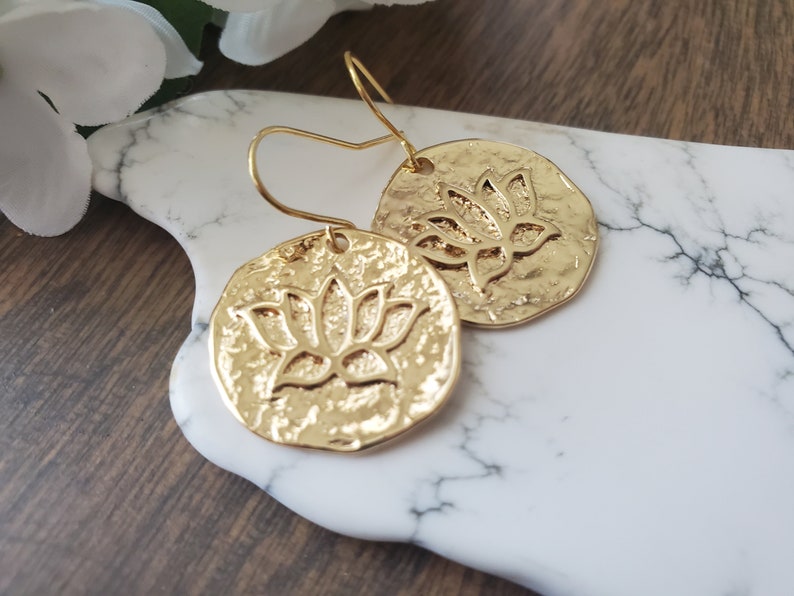 Puede incluir: Pendientes de color dorado con un dise&ntilde;o de flor de loto. Los pendientes son redondos y tienen una textura martillada.