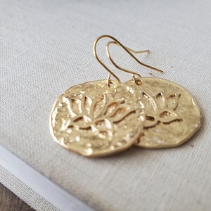 Puede incluir: Pendientes de tono dorado con un dise&ntilde;o circular texturizado que presenta un motivo de flor de loto.