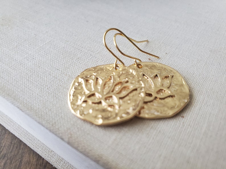 Puede incluir: Pendientes de tono dorado con un dise&ntilde;o circular texturizado que presenta un motivo de flor de loto.