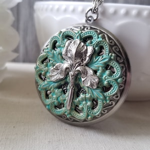 以下が含まれることがあります： ターコイズブルーとシルバーのフローラルデザインが施されたシルバーのロケットネックレス。ロケットには、複雑なディテールが施されたシルバーのアイリスの花が描かれています。