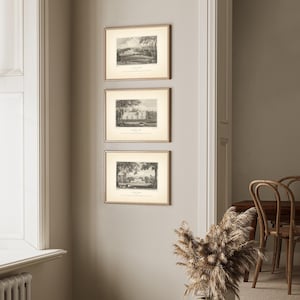 May include: Three framed landscape prints in a neutral-toned room. Each print features a black and white illustration of a scenic view, set within a gold-colored frame. The prints are hung vertically on a light gray wall, creating a classic aesthetic.