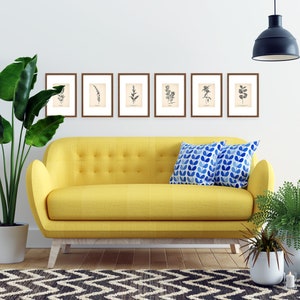 K&ouml;nnte beinhalten: Ein gelbes Sofa mit zwei blauen und wei&szlig;en Kissen steht in einem Wohnzimmer mit einem schwarz-wei&szlig;en Teppich, einer Topfpflanze und einer H&auml;ngelampe. Sechs gerahmte botanische Drucke h&auml;ngen an der Wand &uuml;ber dem Sofa.
