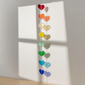Op de afbeelding: Een decoratieve hartvormige zonnevanger in regenboogkleuren, waaronder rood, oranje, geel, groen, blauw en paars. De harten hangen aan een zilveren ketting. Het item wordt gepresenteerd tegen een witte muur.