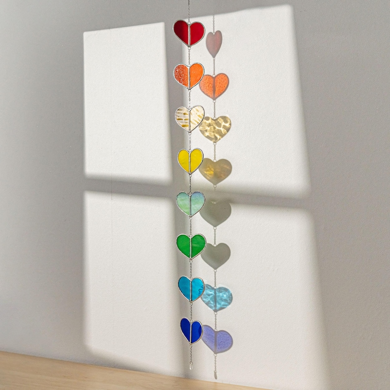 Op de afbeelding: Een decoratieve hartvormige zonnevanger in regenboogkleuren, waaronder rood, oranje, geel, groen, blauw en paars. De harten hangen aan een zilveren ketting. Het item wordt gepresenteerd tegen een witte muur.